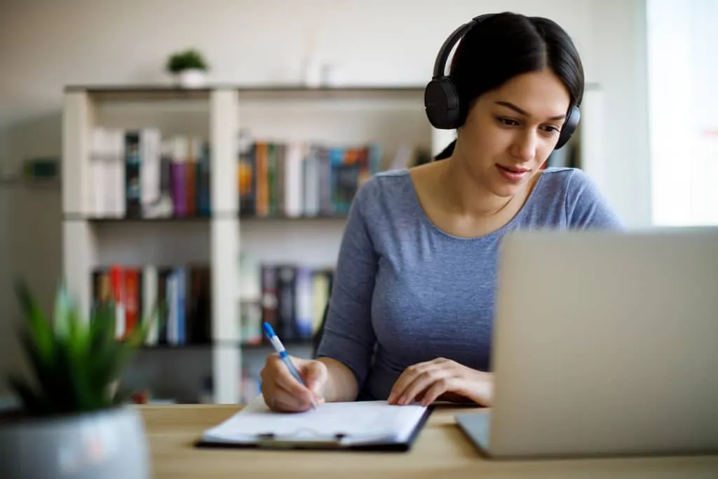 woman wearing headphones, taking notes on clipboard from laptop.