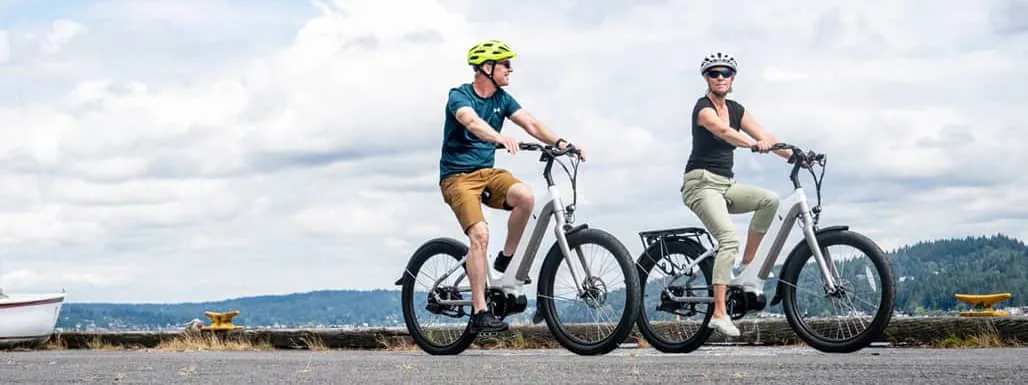 Man and woman riding e-bikes next to boat.